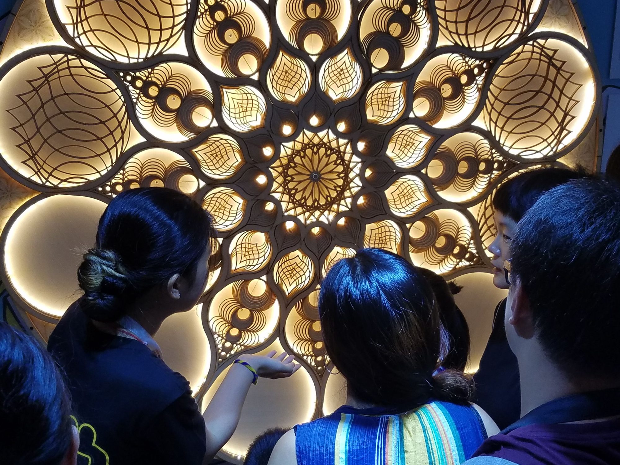 Illuminated laser-cut wood mandala sculpture viewed by a small crowd from behind
