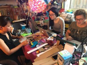 Three people crafting at a table under a lit paper lantern