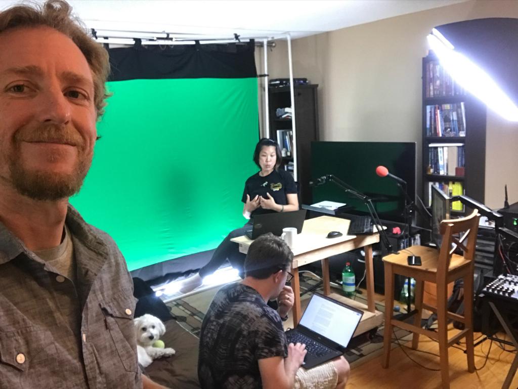 A man takes a selfie with teammates recording a livestream in front of a green screen
