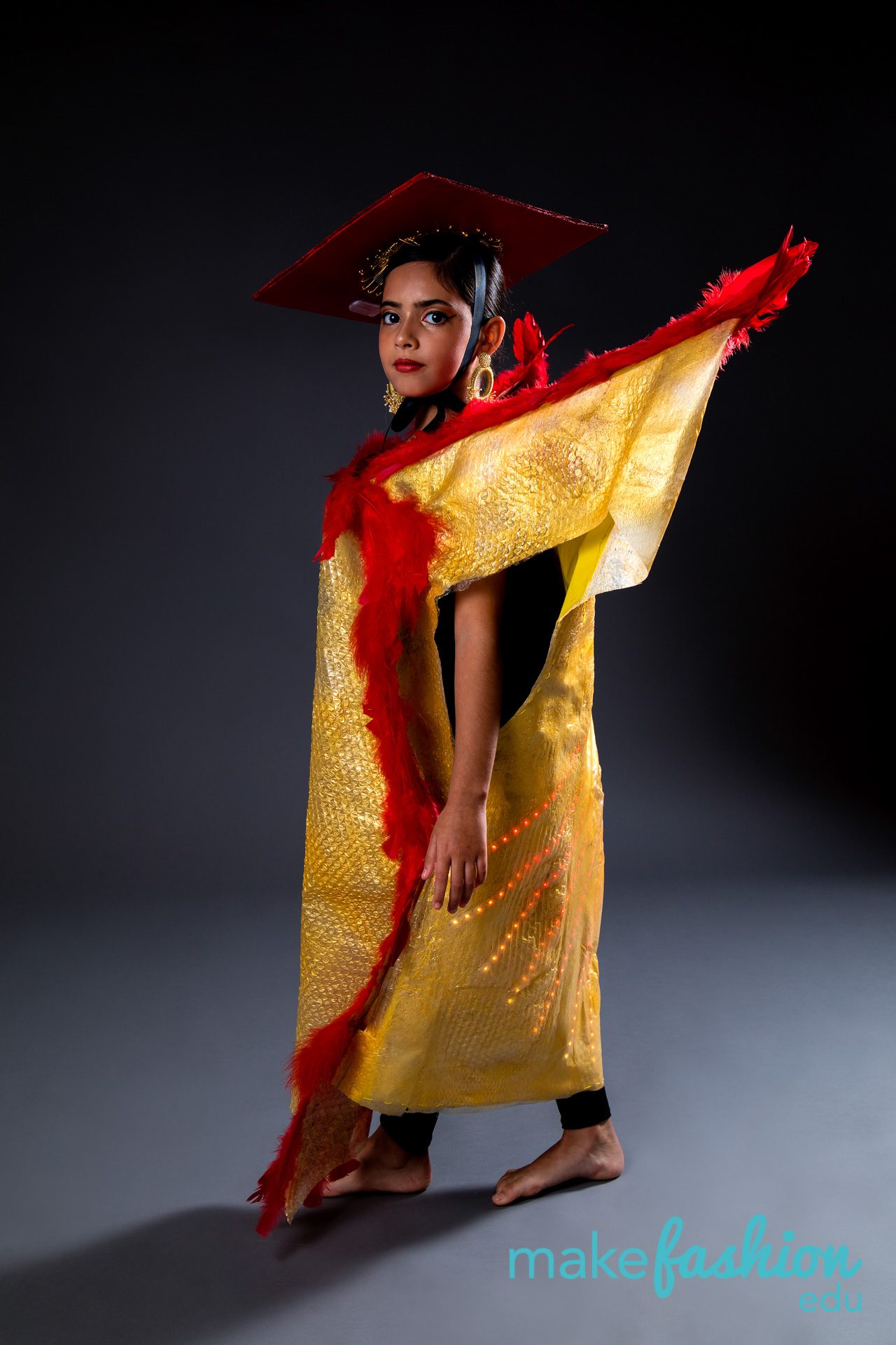 Student model in gold textured dress with red feather trim and a red square hat