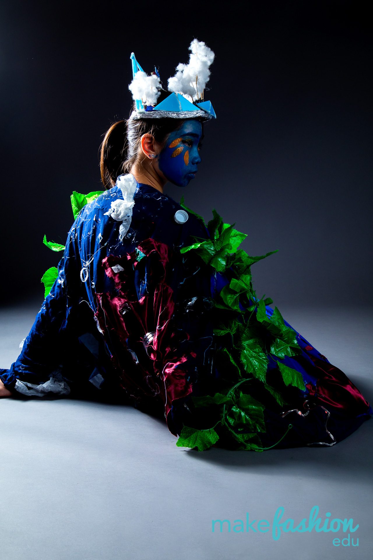 Student model with blue face makeup, cloud headpiece, and dark cape covered in green leaves