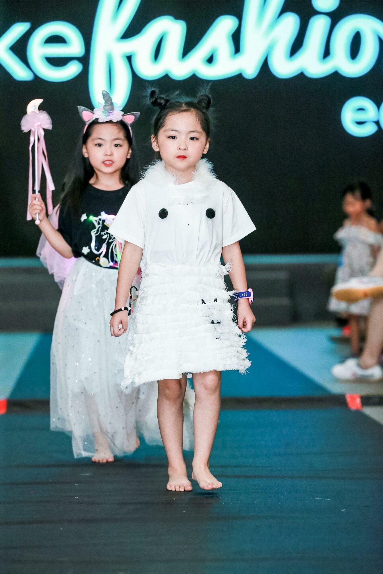 Two students on runway, one in white fringed dress, other with unicorn ears and wand