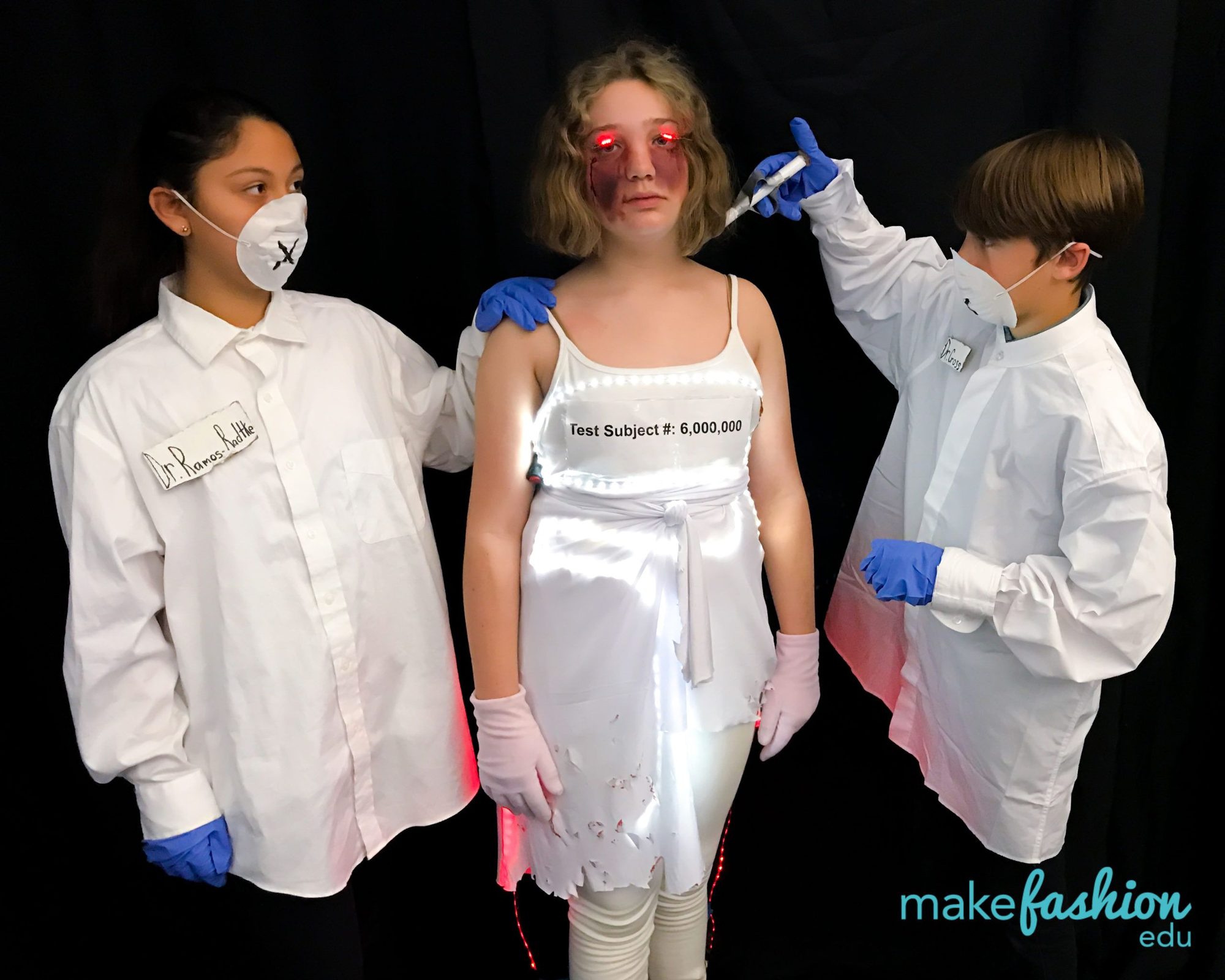 Three students in lab coats beside a glowing LED test-subject dress
